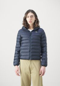 Marinblå puffjacka med huva, som har kviltade sektioner, frontdragkedja och broderad logotyp på vänster bröst. Lätt och strukturerad tyg.