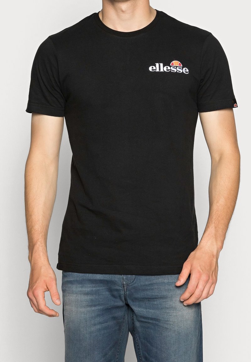 Man i svart ellesse-logot-shirt och blå jeans, står med armarna avslappnat vid sidorna mot en enkel bakgrund.