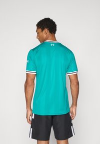 Turkis atletisk drakt med strukturert overflate, med hvitstripet krage og detaljer. Bakfra vises diskret merke på nakken.