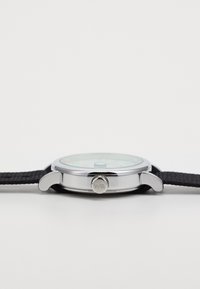 Orologio in acciaio inossidabile con quadrante rotondo verde, cinturino in tessuto nero e una corona con bordo testurizzato. Tra i dettagli ci sono lettere incise sulla corona.