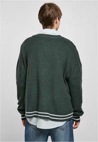 Mørkegrønn strikkegenser med løs passform, med hvite striper på kanten og ermene. Brukt over en lyseblå skjorte med krage.