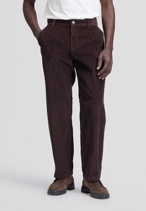 Homme portant un pantalon en velours côtelé marron foncé, des chaussures en suède marron et une chemise blanche, les mains dans les poches, debout sur un fond gris clair.