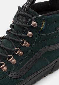 Mørkegrønn høy topp sneaker laget av semsket skinn og tekstil med bronse øyelet, svarte detaljer og en strukturert gummisåle. Har HydroGuard-merke.