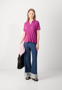 Fuksia lyhythihainen toppi, jossa on kaulus ja nappikiinnitys, yhdistettynä leveälahkeisiin sinisiin farkkuihin, joissa on purkautuva helma ja harmaat yksityiskohdat. Musta kassi.