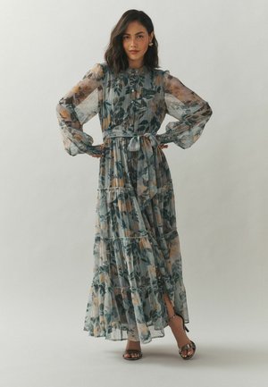 Woman standing with hands on hips wearing a long, sheer floral dress with puffed sleeves and ankle-strap heels on a plain background.