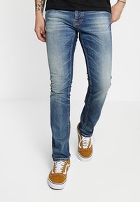 Blå denimjeans med slim passform, lätt blekning och whiskering, parat med bruna mockasneakers med vita snören och gummisulor.