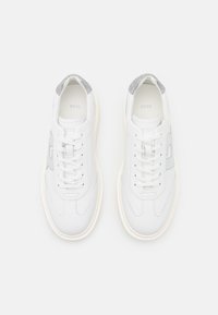 Bílé tenisky se hladkým koženým svrškem, šedými textilními akcenty na patě a bílou mezipodešví. Obsahují šňůrky a detail s logem na boku.