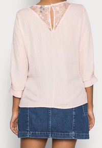Helle rosa Bluse mit drei Viertelärmeln, Spitzenbesatz am Rückenausschnitt und aufgerollten Bündchen. Kombiniert mit einem Jeansrock.