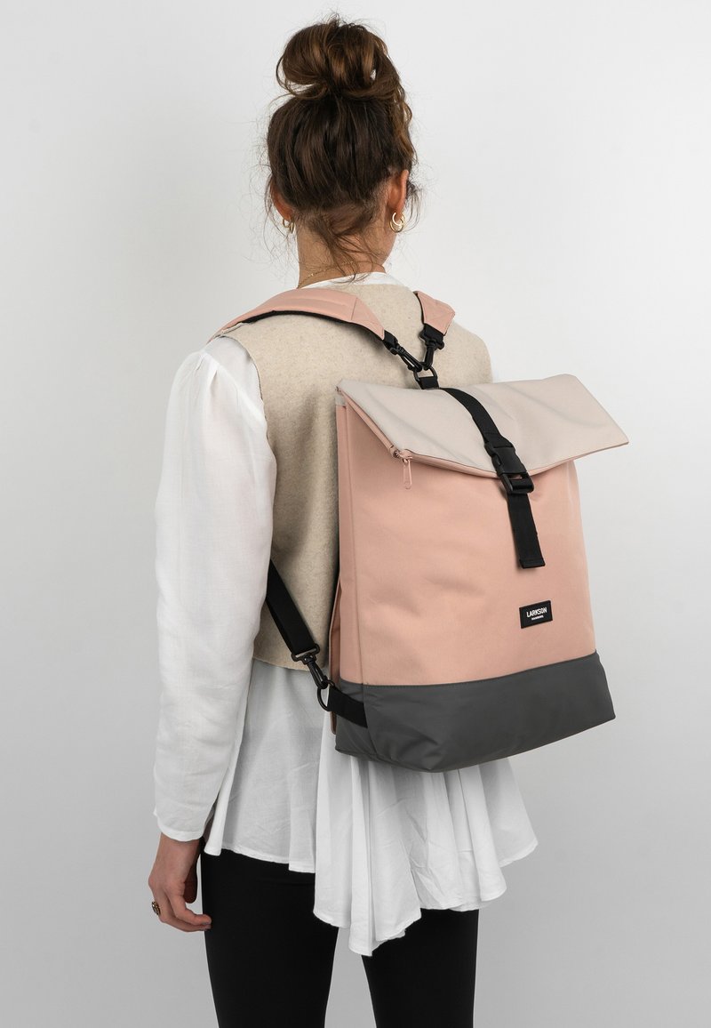Rózsaszín és szürke hátizsák, feltekerhető dizájnnal, szövetből készült. Fekete hardverrel és állítható pántokkal rendelkezik, egy modellen bemutatva.