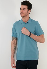 Tricou polo albastru cu guler clasic, mâneci scurte și închidere cu trei butoane, fabricat dintr-un material neted. Purtați cu pantaloni închiși la culoare.