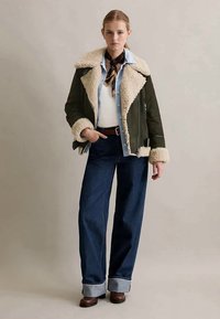 Mulher vestindo uma jaqueta de pele sintética verde-oliva, camisa azul clara, suéter branco, jeans largos escuros, botas castanhas e um lenço de seda no pescoço, em pé com a mão no bolso.
