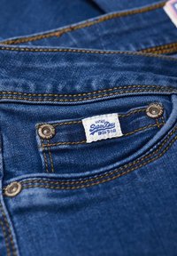 Tela de mezclilla azul con costuras doradas, que presenta un bolsillo y una etiqueta blanca que dice "Superdry". Botón a presión de metal plateado visible.