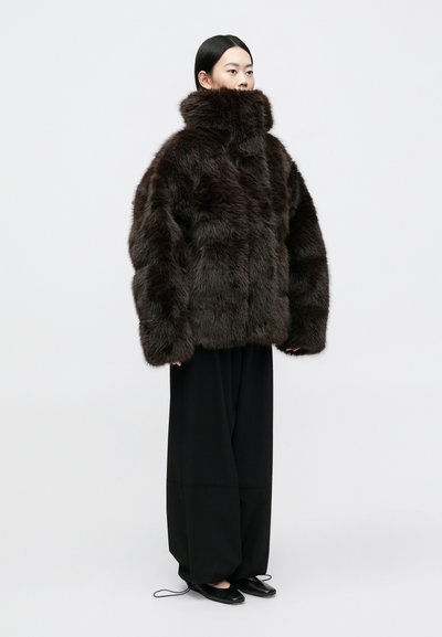 Ruda dirbtinė kailinė striukė su aukštu apykakle, laisvu kirpimu ir tekstūrine paviršiumi; derinama su plačiomis juodomis kelnėmis ir lygiais juodais batais.