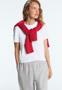 Žena oblečená v bielom tričku a sivých teplákových nohaviciach s červeným svetrom prehodeným cez plecia, pozerajúca sa do strany, ruky v vreckách.