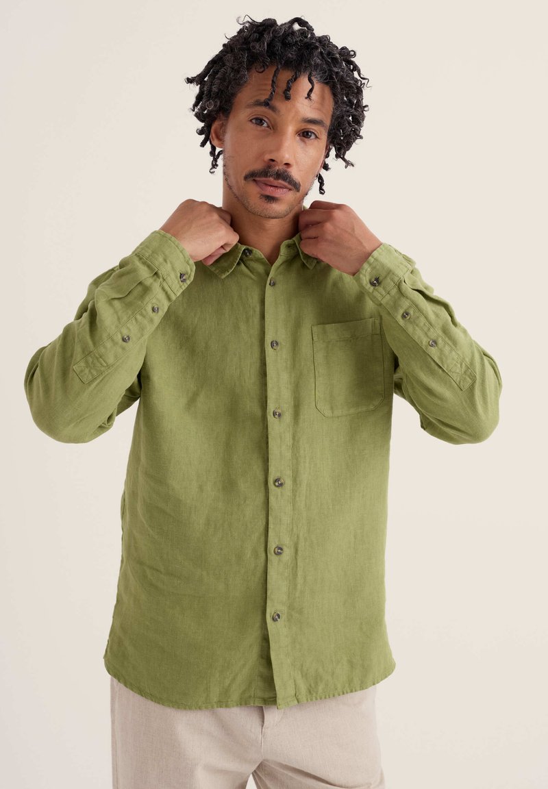 Homme ajustant le col d'une chemise boutonnée à manches longues de couleur vert olive, debout devant un fond clair uni.