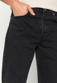 Jeans de mezclilla negra con un corte clásico, que presentan un diseño de cinco bolsillos, costuras visibles y una textura sutil. Mano descansando en el bolsillo.