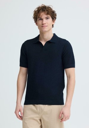 Junger Mann mit lockigem Haar, der ein kurzärmeliges, strukturiertes schwarzes Poloshirt und beige Hosen trägt, stehend vor einem einfarbigen hellen Hintergrund.