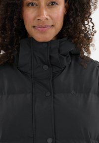 Schwarze gepolsterte Jacke mit hohem Kragen, aus glattem Stoff, großen Knöpfen und einem dezenten Logo auf der Brust.