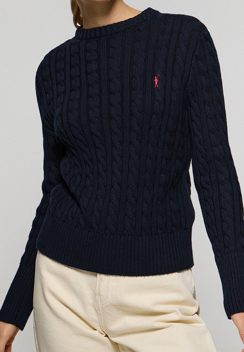 Pull bleu marine en maille torsadée avec logo rouge brodé sur la poitrine, porté avec un pantalon beige taille haute.