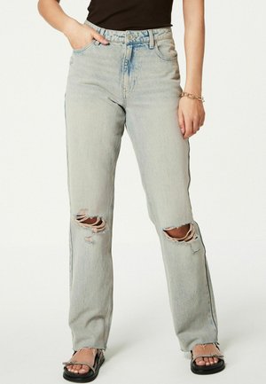 Kobieta w jasnoniebieskich, prostych jeansach z dziurami, czarnym bezrękawniku, czarnych sandałach i złotej bransoletce na lewej ręce.