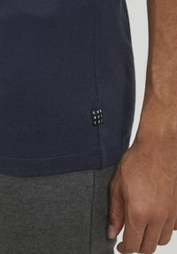 Námořnická modrá bavlněná tričko s krátkým rukávem, s malou černou značkou na lemu. Jemná textura, kombinované s šedými látkovými kalhotami.