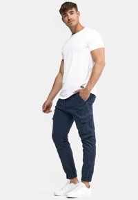 Camiseta blanca de manga corta, pantalones cargo azul marino con puños elásticos, múltiples bolsillos y zapatillas blancas. Diseño simple y funcional.