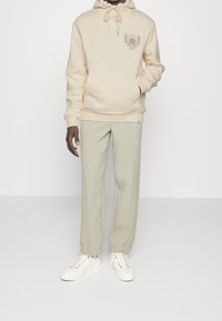 Sudadera beige con capucha ajustable y emblema, combinada con pantalones jogger verde claro. Zapatillas blancas con ojales multicolores completan el conjunto.