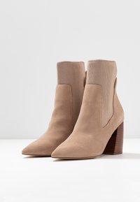 Beżowe zamszowe botki za kostkę z żebrowanym dzianinowym wierzchem, spiczastym noskiem i brązowym drewnianym obcasem w kształcie klocka. Posiadają elastyczne boczne panele, co ułatwia zakładanie.
