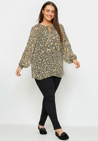 Blusa beige con stampa leopardata nera, maniche lunghe e scollatura a chiave, abbinata a pantaloni neri slim-fit e mocassini neri.