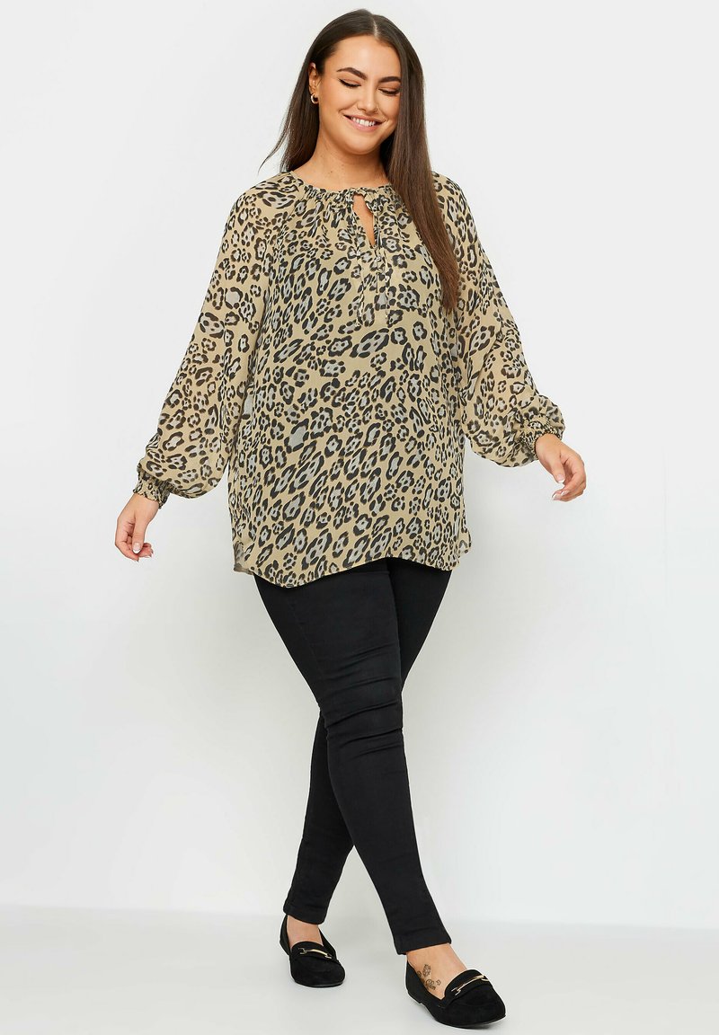 Blusa beige con stampa leopardata nera, maniche lunghe e scollatura a chiave, abbinata a pantaloni neri slim-fit e mocassini neri.