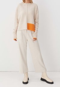 Beżowy sweter dzianinowy z pomarańczowym ściągaczem, w oversize'owym fasonie, z długimi rękawami. Połączony z kremowymi spodniami z szerokimi nogawkami i beżowymi botkami z czarnymi podeszwami.