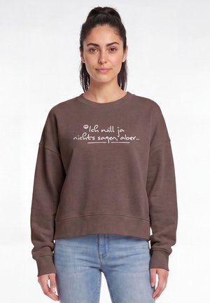 Vrouw met donker haar in een staart, draagt een bruin sweatshirt met Duitse tekst en lichtblauwe jeans tegen een witte achtergrond.