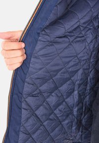Una mano tiene aperto un giubbotto trapuntato di colore blu navy, mostrando l'imbottitura a diamante e un bordo della zip a righe multicolori.