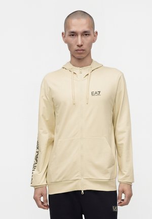 Homme à la tête rasée portant un sweat à capuche beige zippé avec le logo EA7 sur la poitrine et le texte Emilio Armani sur la manche gauche, associé à un pantalon noir.