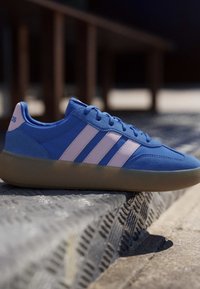 Blue Adidas sneaker with white stripes and a gum sole resting on a textured metal surface outdoors in sunlight.