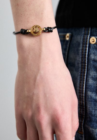 Bracciale in pelle nera con chiusura rotonda in oro a superficie texture, indossato su un polso su uno sfondo chiaro.