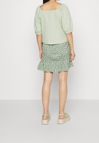 Top verde chiaro a maniche a sbuffo con scollo quadrato, abbinato a una gonna a balze e a fantasia in diverse tonalità di verde. Sandali semplici e piatti completano il look.