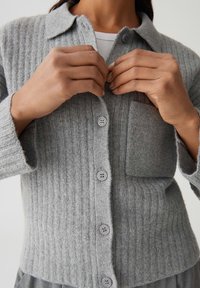 Cardigan gris à côtes avec col, comportant une poche avant, des boutons et un tissu texturé, montré en train d'être boutonné par des mains.