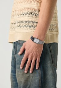 Metallisk klokke med rektangulært urskive, sølvfarget og integrert leddarmbånd. Modellen viser intrikat strikket topp og denimjeans.