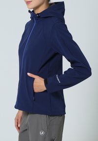 Chaqueta softshell azul marino con capucha, que cuenta con un cierre frontal, bolsillos laterales y puños elásticos en contraste. Textura de tela suave.