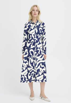 Mujer vistiendo un vestido midi abotonado de manga larga con estampado floral abstracto en azul marino y blanco, acompañado de tacones slingback blancos con punta puntiaguda.