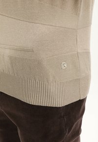 Beez värviga kootud sviiter, millel on tekstuurilised mustrid ja ribaline äär, ning väike logo detail küljes.