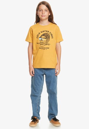 Camiseta de algodón amarilla con un estampado gráfico que presenta el texto "COLD WATER VIBES" y un diseño de calavera, combinada con unos jeans de mezclilla azules y zapatos marrones.