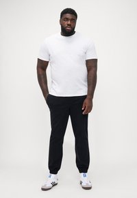Hvid t-shirt med korte ærmer, sorte joggerbukser med elastiske cuffs, og hvide sneakers med blå detaljer, stående mod en ensfarvet baggrund.