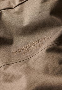 Brunt tekstureret stof med et præget "SUPERDRY NYC SPORTSWEAR" logo i beige. Materialet virker let og afslappet.