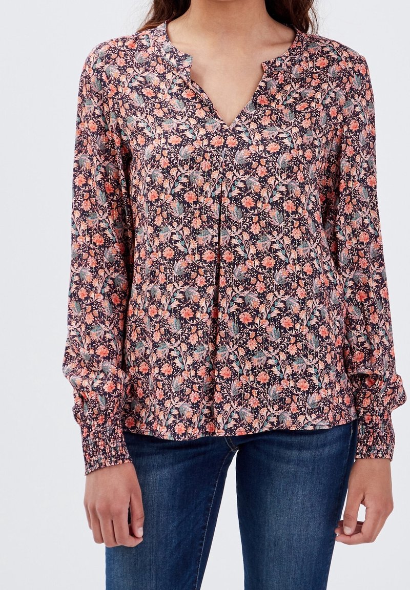 Femme portant un chemisier à manches longues à fleurs roses, oranges et vertes, associé à un jean bleu foncé, debout les bras détendus.