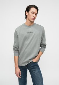 Γκρι μακρυμάνικη μπλούζα με στρογγυλή λαιμόκοψη και λογότυπο Calvin Klein στο στήθος, συνδυασμένη με μπλε τζιν. Απλό, λείο ύφασμα και άνετη εφαρμογή.