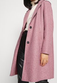 Nainen, joka pukeutuu nappilangattomaan vaaleanpunaiseen villakangastakkiin, jonka alla on kerma värinen poolopaita ja mustat nahkahousut, yksinkertaista valkoista taustaa vasten.