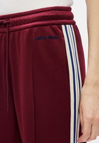 Βαμβακερό παντελόνι φόρμας μπορντό με μπλε και λευκές ρίγες στο πλάι, με κορδόνι στη μέση, μικρό λογότυπο "calvin klein" κοντά στη τσέπη, φορεμένο από άτομο με το χέρι ορατό.