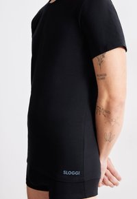 Černé tričko SLOGGI s kulatým výstřihem, krátkými rukávy a slim fit střih. Disponuje hladkým materiálem a modrou logovou aplikací blízko lemu.